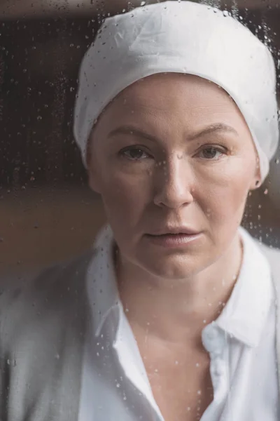 Vista de cerca de la mujer madura enferma en el pañuelo mirando a la cámara a través de la ventana con gotas de lluvia - foto de stock
