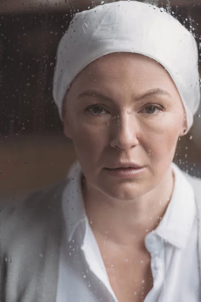 Vue rapprochée de femme mature malade en mouchoir regardant la caméra à travers la fenêtre avec des gouttes de pluie — Photo de stock