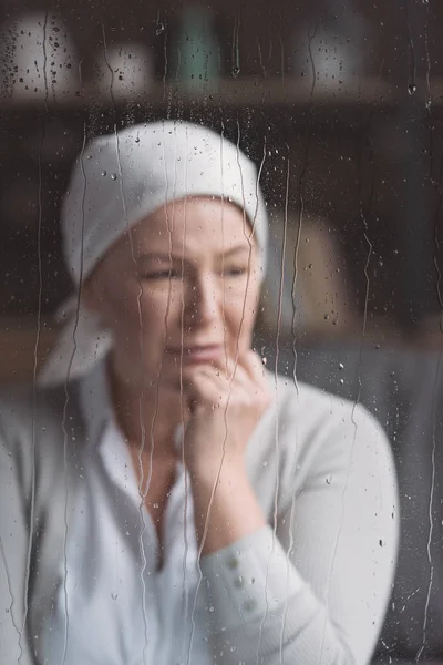 Mujer madura enferma en pañuelo llorando detrás de la ventana con gotas de lluvia - foto de stock