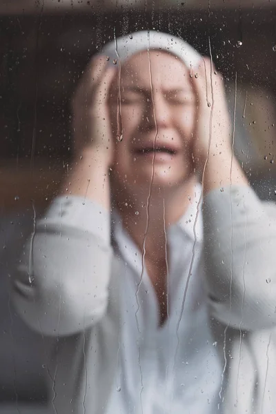 Mujer madura enferma en pañuelo llorando detrás de la ventana con gotas de lluvia - foto de stock