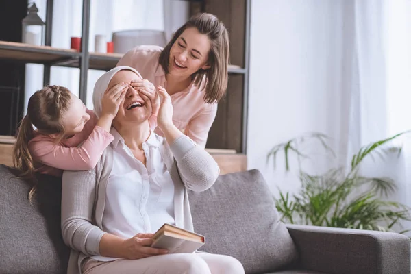 Mother and daughter visiting smiling sick grandmother in kerchief reading book at home — Stock Photo