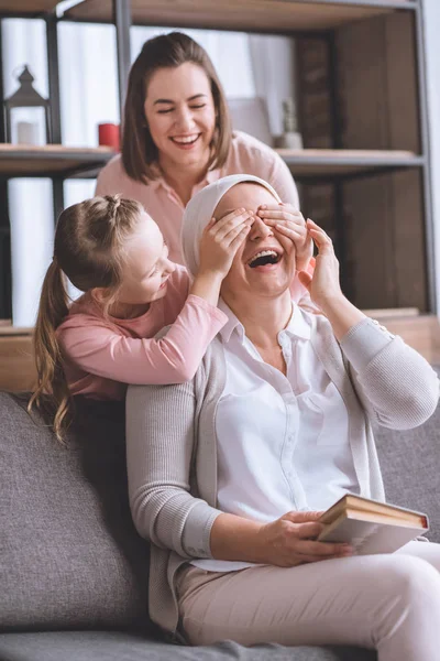 Happy mother and daughter visiting sick smiling grandmother in kerchief reading book at home — Stock Photo