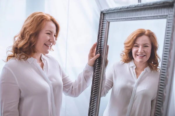 Belle femme mûre souriante debout près du miroir et regardant la réflexion — Photo de stock