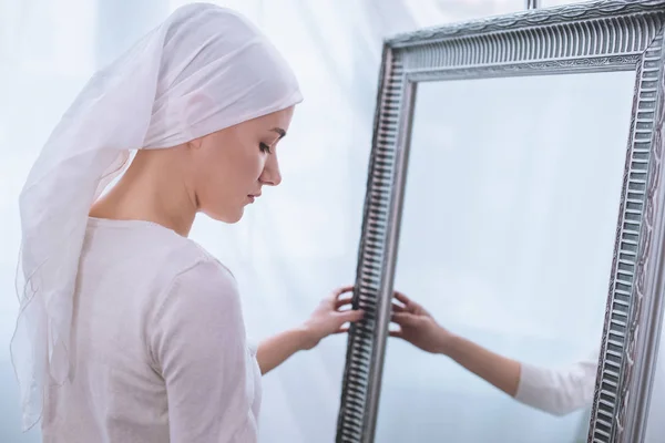 Вид сбоку на молодую больную женщину в платке, стоящую у зеркала, концепция рака — стоковое фото