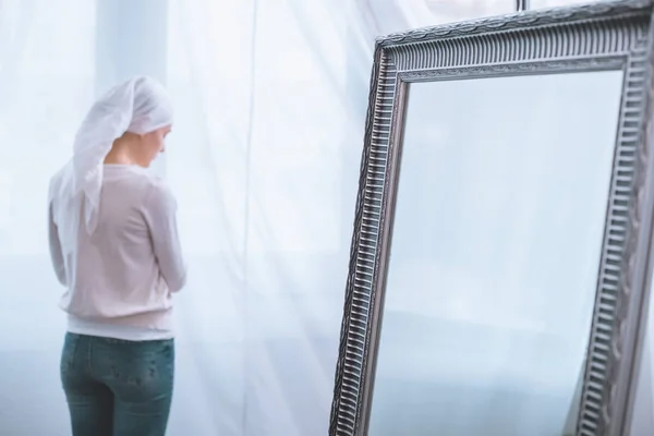 Back view of young sick woman in kerchief standing near mirror, cancer concept — Stock Photo