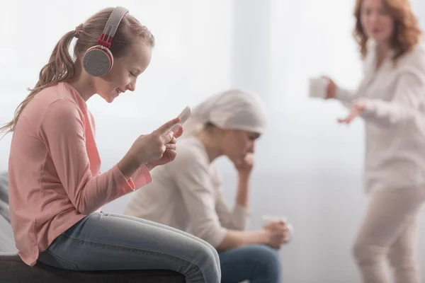 Niño en auriculares usando tableta digital mientras madre enferma y abuela hablando detrás - foto de stock