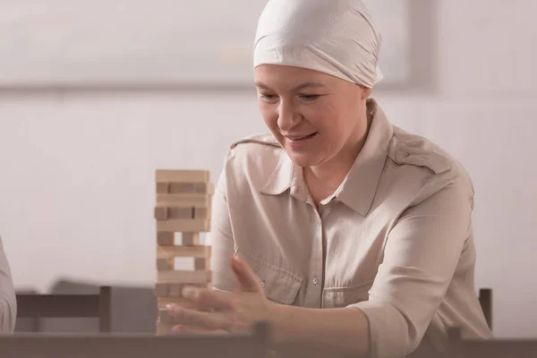 Mujer madura sonriente enferma en torre de construcción de pañuelo de bloques de madera en casa - foto de stock