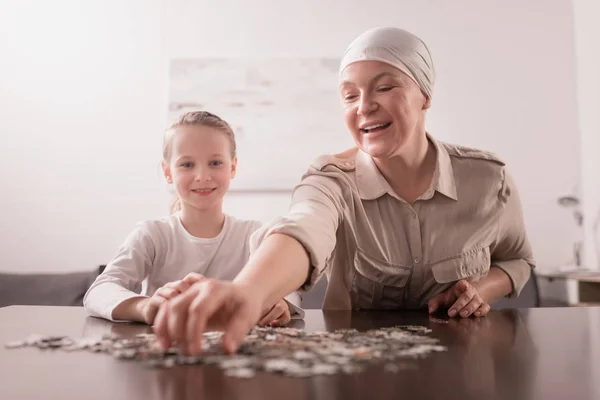 Smiling kid with sick grandmother in kerchief playing with jigsaw puzzle together — Stock Photo
