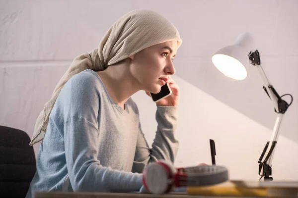 Side view of young woman in kerchief talking by smartphone and taking notes — Stock Photo