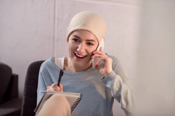 Smiling young woman in kerchief talking by smartphone and taking notes — Stock Photo