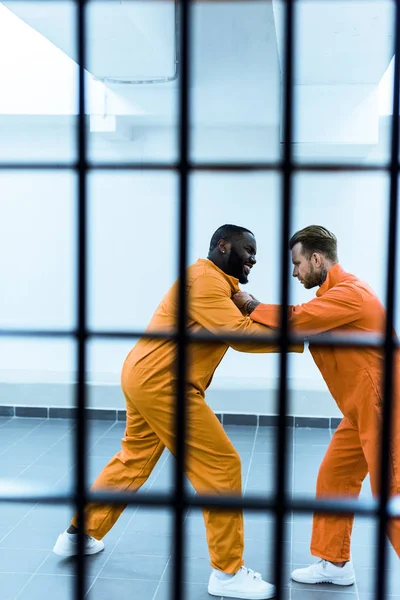 Vista lateral de prisioneros multiculturales amenazándose entre sí tras las rejas de las prisiones - foto de stock