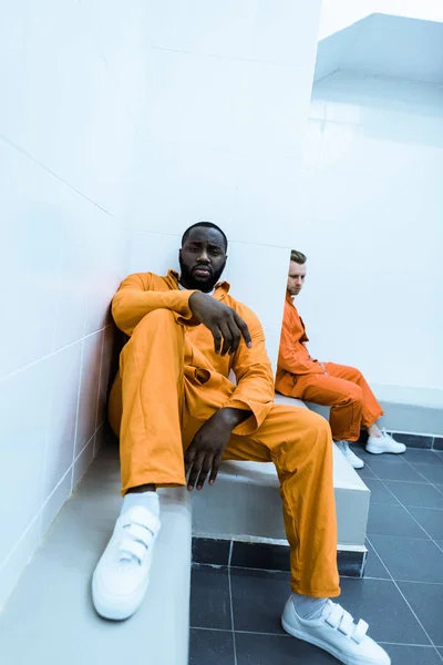 Presos multiculturales sentados en bancos en la celda de la prisión - foto de stock