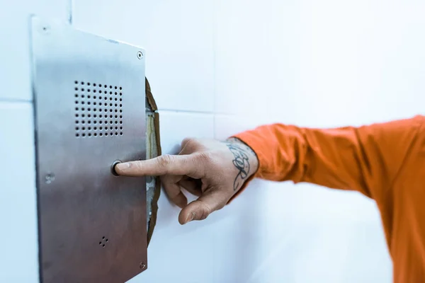 Cropped image of tattooed prisoner in orange uniform pressing button in prison cell — Stock Photo