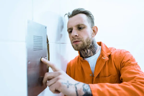 Tätowierter Häftling in orangefarbener Uniform drückt in Gefängniszelle Knopf — Stockfoto