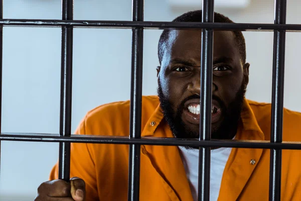 Prisionero afroamericano enojado sosteniendo barras de prisión - foto de stock