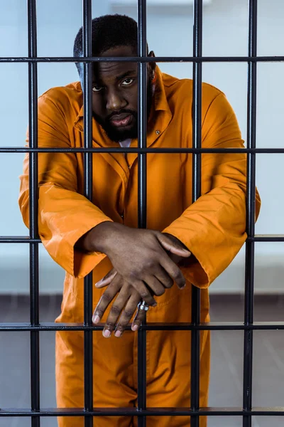 Prisionero afroamericano en uniforme parado detrás de las rejas de la prisión - foto de stock