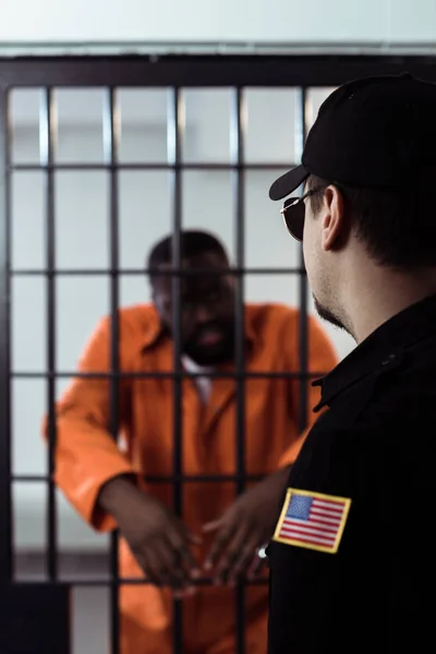 Guardia de seguridad de pie cerca de bares de la prisión y mirando a un prisionero afroamericano - foto de stock