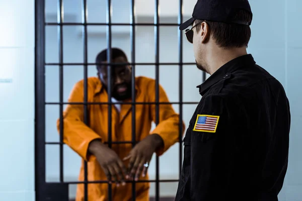 Oficial de la prisión de pie cerca de bares de la prisión y mirando a un prisionero afroamericano - foto de stock
