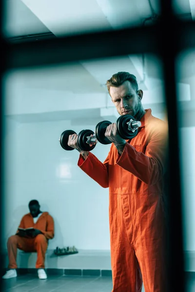 Prisoner training with dumbbells in prison room behind prison bars — Stock Photo