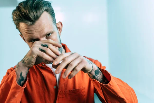 Prisionero con tatuajes cubriendo la cara con las manos y mirando a la cámara - foto de stock