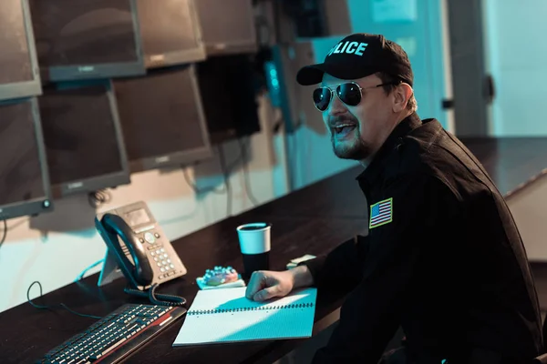 Rindo guarda prisional sentado à mesa de trabalho e monitorando pessoas na prisão — Fotografia de Stock