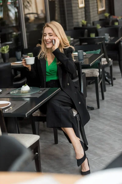 Mulher sorrindo com xícara de café falando no smartphone no café — Fotografia de Stock
