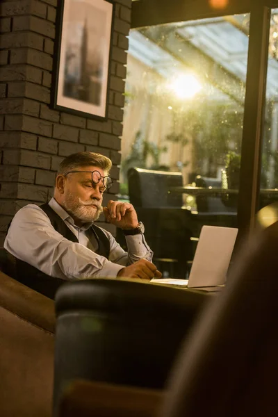 Side view of pensive senior businessman sitting at table with laptop in cafe — Stock Photo