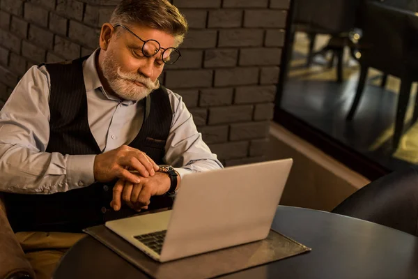 Hombre de negocios senior comprobar el tiempo mientras está sentado en la mesa con el ordenador portátil en la cafetería - foto de stock