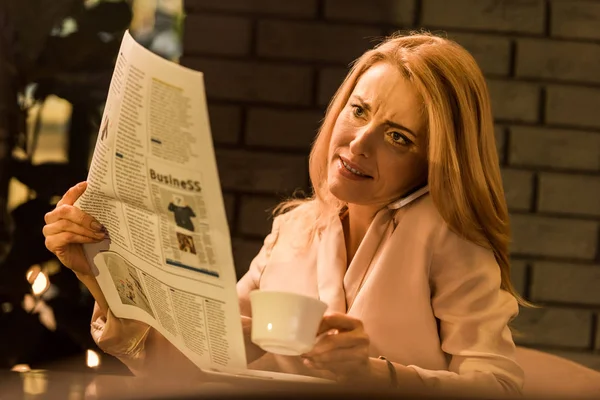 Portrait de femme d'affaires avec tasse de café parler sur smartphone tout en lisant le journal dans le café — Photo de stock