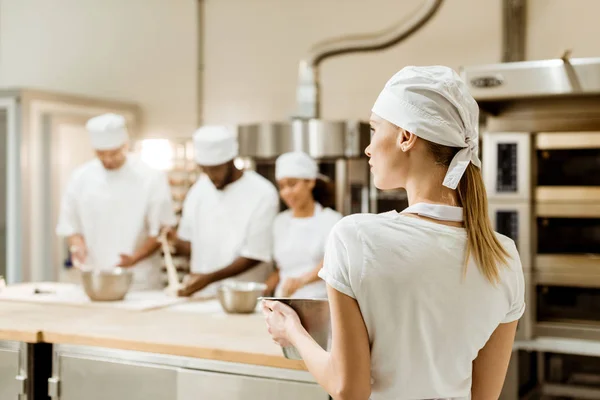 Обратный вид молодой пекарши на рабочем месте с размытыми коллегами, работающими на заднем плане — стоковое фото