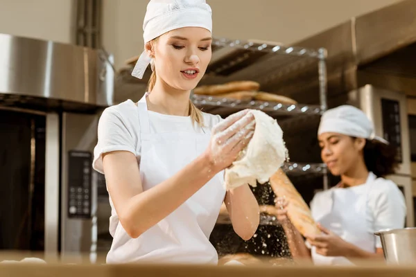 Young female baker kneading dough at baking manufacture while her colleague working blurred on background — Stock Photo