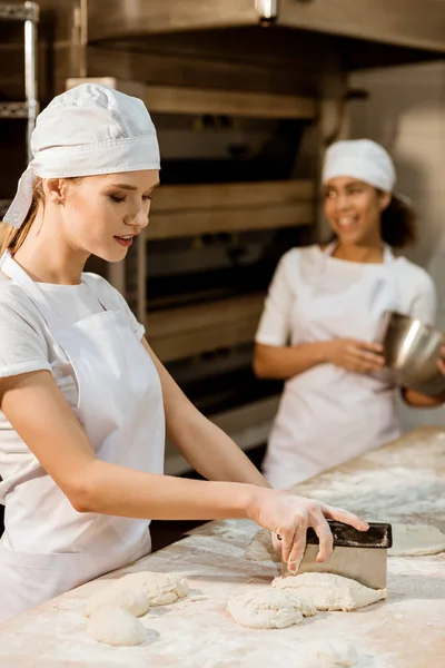 Young female baker cutting dough with dough knife at baking manufacture while her colleagues working blurred on background — Stock Photo