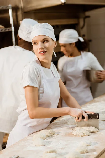 Atraente mulher padeiro corte massa com faca de massa na fabricação de cozimento, enquanto seus colegas de trabalho desfocado no fundo — Fotografia de Stock