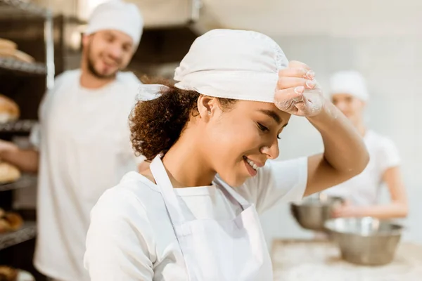 Счастливая и измученная пекарь работает на выпечке, в то время как ее коллеги работают размыто на заднем плане — стоковое фото