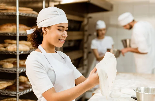Американская пекарь-женщина смешивает тесто при производстве выпечки, в то время как ее коллеги размываются на заднем плане — стоковое фото