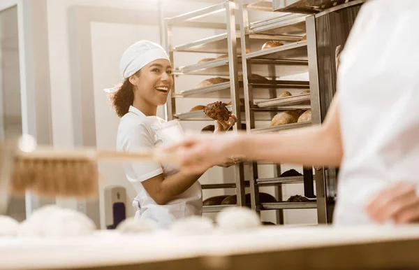 Jeune boulangère parlant à un collègue à la fabrication de cuisson pendant qu'elle nettoie le lieu de travail de la farine — Photo de stock