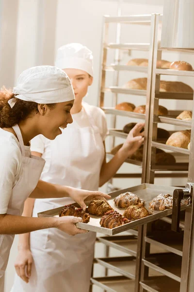 Panaderos femeninos jóvenes poniendo pastelería fresca en los estantes en la fabricación de hornear - foto de stock
