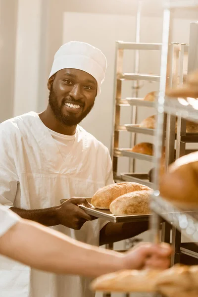 Smiling african american baker holding loaves of bread on tray at baking manufacture — Stock Photo
