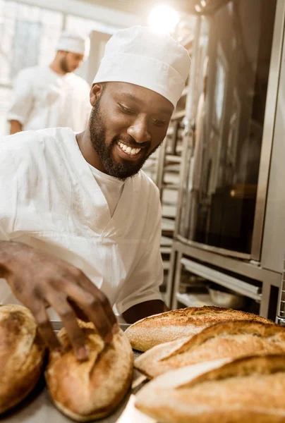 Smiling african american baker with tray of fresh loaves of bread on baking manufacture — Stock Photo