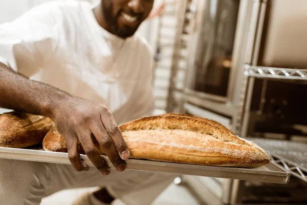 Tiro recortado de panadero afroamericano con bandeja de panes frescos en la fabricación de hornear - foto de stock