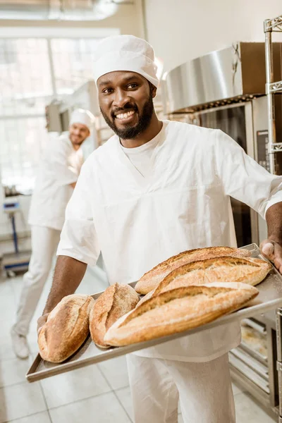African american baker taking bread loaves from oven at baking manufacture — Stock Photo