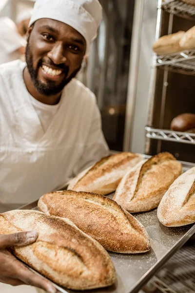 Smiling handsome african american baker with tray of fresh loaves of bread on baking manufacture — Stock Photo