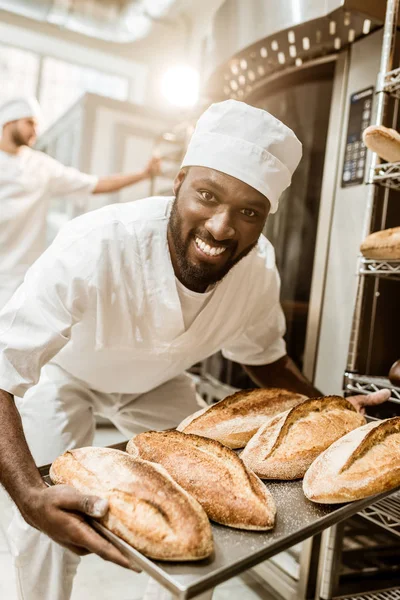Happy african american baker taking bread loaves from oven at baking manufacture — Stock Photo
