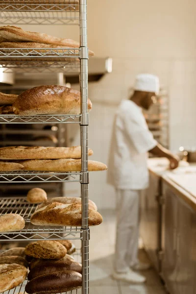 Африканский американский пекарь готовит сырое тесто на рабочем месте с полками свежего хлеба на переднем плане — стоковое фото