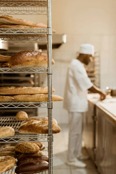 African american baker preparing raw dough at workplace with shelves of fresh bread on foreground — Stock Photo