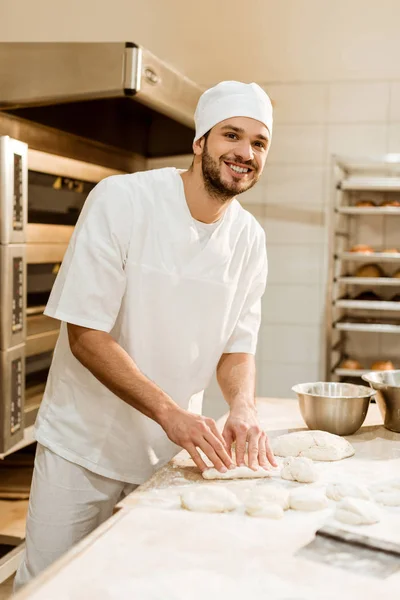 Sorridente giovane panettiere preparare pasta cruda sul posto di lavoro sulla fabbricazione di cottura — Foto stock