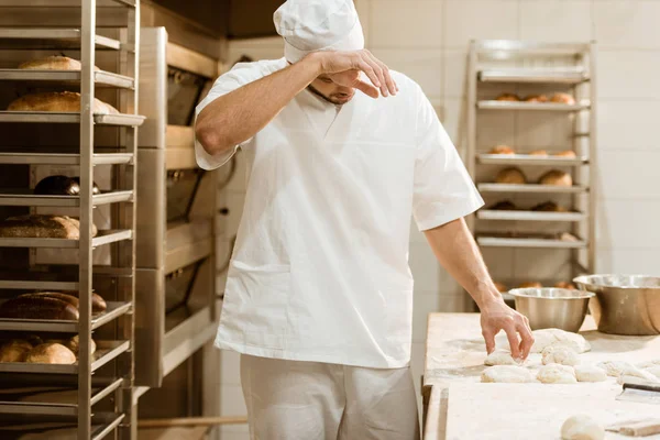 Erschöpfter Bäcker arbeitet mit rohem Teig am Backwerk und wischt Schweiß ab — Stockfoto