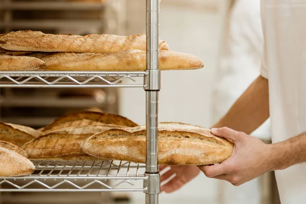 Cropped shot of handsome baker putting fresh bread on shelf at baking manufacture — Stock Photo