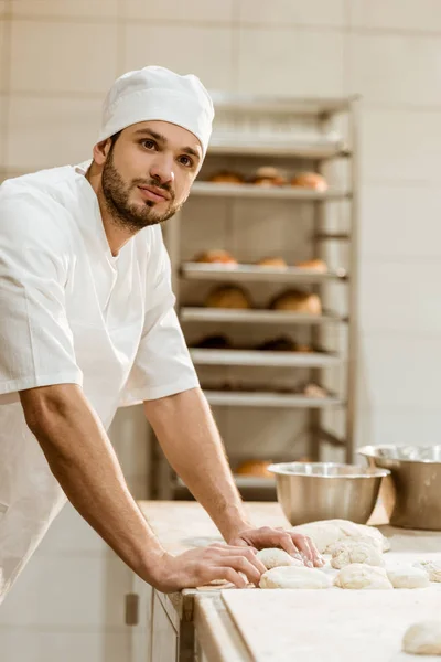 Красивый молодой пекарь с сырым тестом на рабочем месте по производству выпечки — стоковое фото