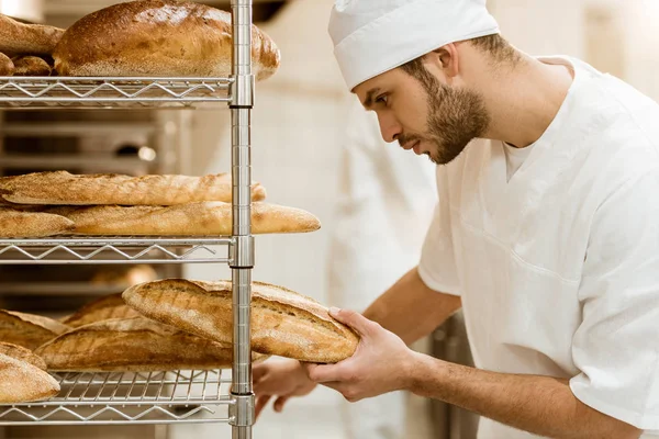 Side view of handsome baker putting fresh bread on shelf at baking manufacture — Stock Photo